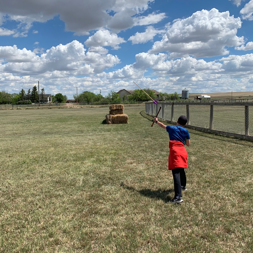 A child  stands in a grassy field holding an atlatl, preparing to throw it towards a stack of hay bales under a cloudy sky.
