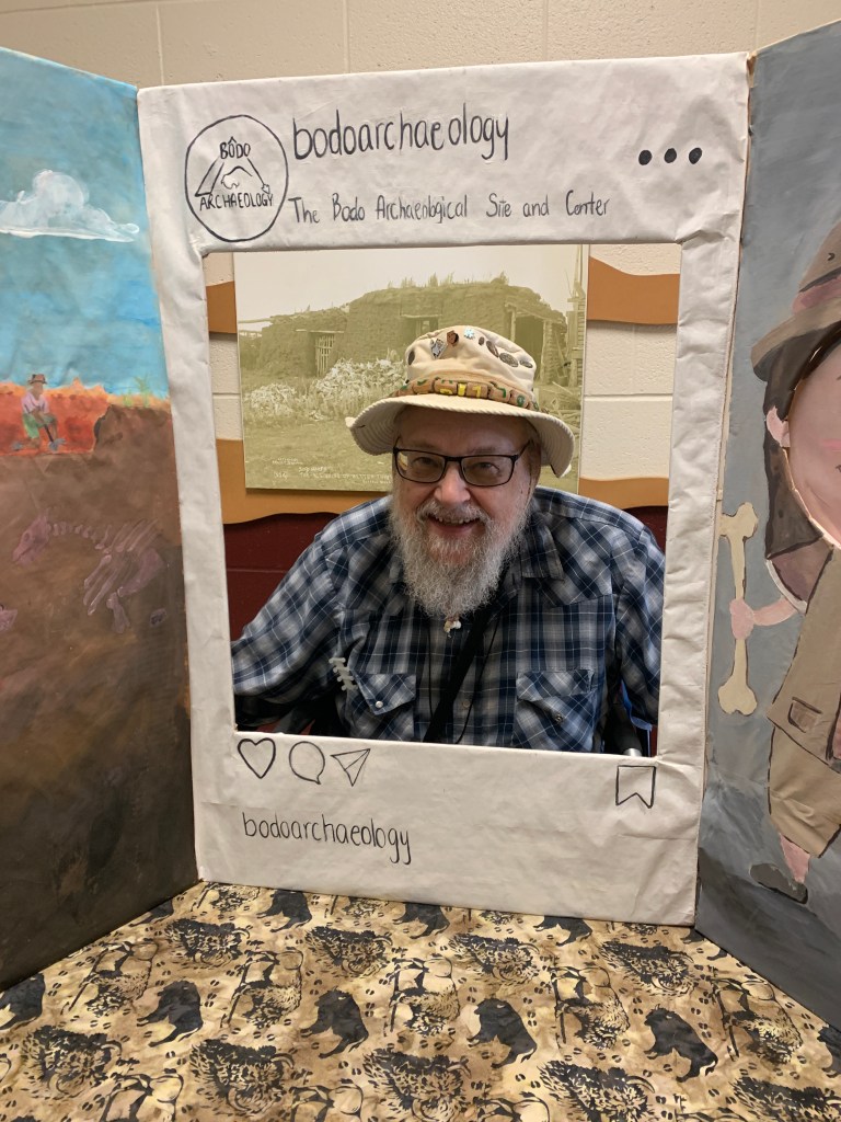 Peter Kirchmeir sits in front of a backdrop showcasing the Bodo Archaeological Site, framed by a decorative structure resembling a social media post.